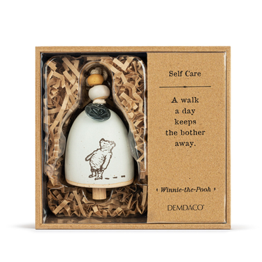 A ceramic bell with a silhouette of Winnie the Pooh, suspended from a beige cord with wooden and ceramic beads. The bell is displayed in a box with a 'Self Care' note and a quote from Winnie the Pooh.