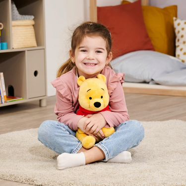 Child holding a Winnie the Pooh plush toy in a cozy room with pillows and shelves.