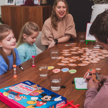 Family playing a board game together at a table with colorful game pieces and a box on the table.